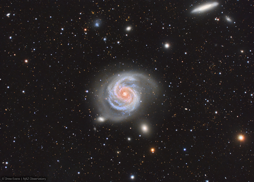 Mitten im Bild leuchtet eine Spiralgalaxie, die direkt von oben zu sehen ist. Ihr Zentrum ist orangefarben mit einem gelben Kern. Darum herum sind Sterne und weitere Galaxien verteilt.